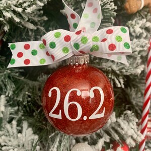 May include: Red glitter Christmas ornament with a white ribbon and white polka dots. The ornament has the number 26.2 written on it in white.