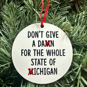 May include: White round Christmas ornament with black text that reads "Don't give a daxn for the whole state of Michigan". The word "daxn" and "Michigan" have red Xs over them.