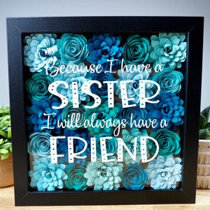 May include: A framed shadow box with a blue and teal paper flower arrangement. The text "Because I have a sister I will always have a friend" is written in white on the glass.