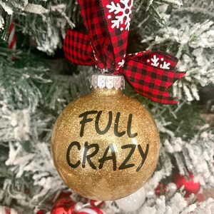 May include: Gold glitter Christmas ornament with black text that reads "FULL CRAZY". The ornament is hanging from a red and black buffalo plaid ribbon with white snowflakes.