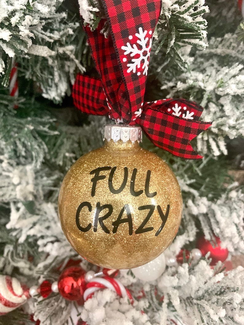 May include: Gold glitter Christmas ornament with black text that reads "FULL CRAZY". The ornament is hanging from a red and black buffalo plaid ribbon with white snowflakes.