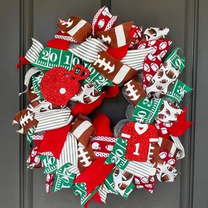 May include: A decorative wreath featuring football-themed ribbons in red, green, white, and brown. The design includes patterns of footballs, field yard lines, and glittery red cutouts of a football helmet and a jersey with the number one.