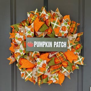 May include: A decorative wreath featuring a mix of orange and green mesh ribbons, patterned fabric ribbons with pumpkin and leaf prints, and a central rectangular dark green sign with the text PUMPKIN PATCH and a small red pumpkin icon.