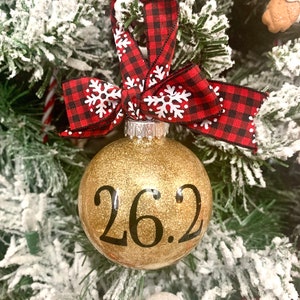 May include: Gold glitter Christmas ornament with black text that reads "26.2". The ornament is decorated with a red and black buffalo plaid ribbon with white snowflakes.