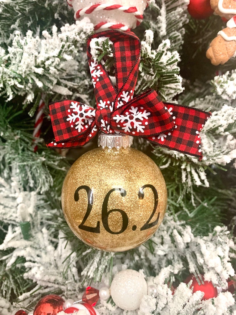 May include: Gold glitter Christmas ornament with black text that reads "26.2". The ornament is decorated with a red and black buffalo plaid ribbon with white snowflakes.