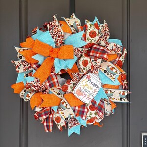 May include: A decorative wreath featuring orange, light blue, and plaid ribbons mixed with patterned fabric showing pumpkins and wheelbarrows. A central sign shaped like a mason jar displays the text Pumpkin Spice & EVERYTHING Nice.