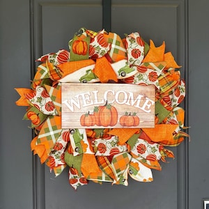 May include: A decorative wreath featuring orange, green, and white ribbons with pumpkin and truck patterns. A rectangular wooden sign in the center displays the word WELCOME in white letters above an illustration of three orange pumpkins.
