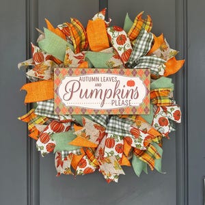 May include: A decorative wreath featuring orange, green, and plaid ribbons interspersed with fabric patterns of pumpkins and autumn leaves. A rectangular sign in the center displays the text "AUTUMN LEAVES AND Pumpkins PLEASE" in brown and orange.