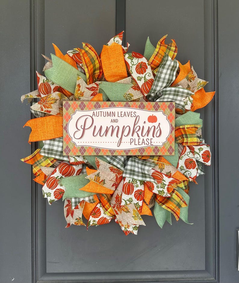 May include: A decorative wreath featuring orange, green, and plaid ribbons interspersed with fabric patterns of pumpkins and autumn leaves. A rectangular sign in the center displays the text "AUTUMN LEAVES AND Pumpkins PLEASE" in brown and orange.