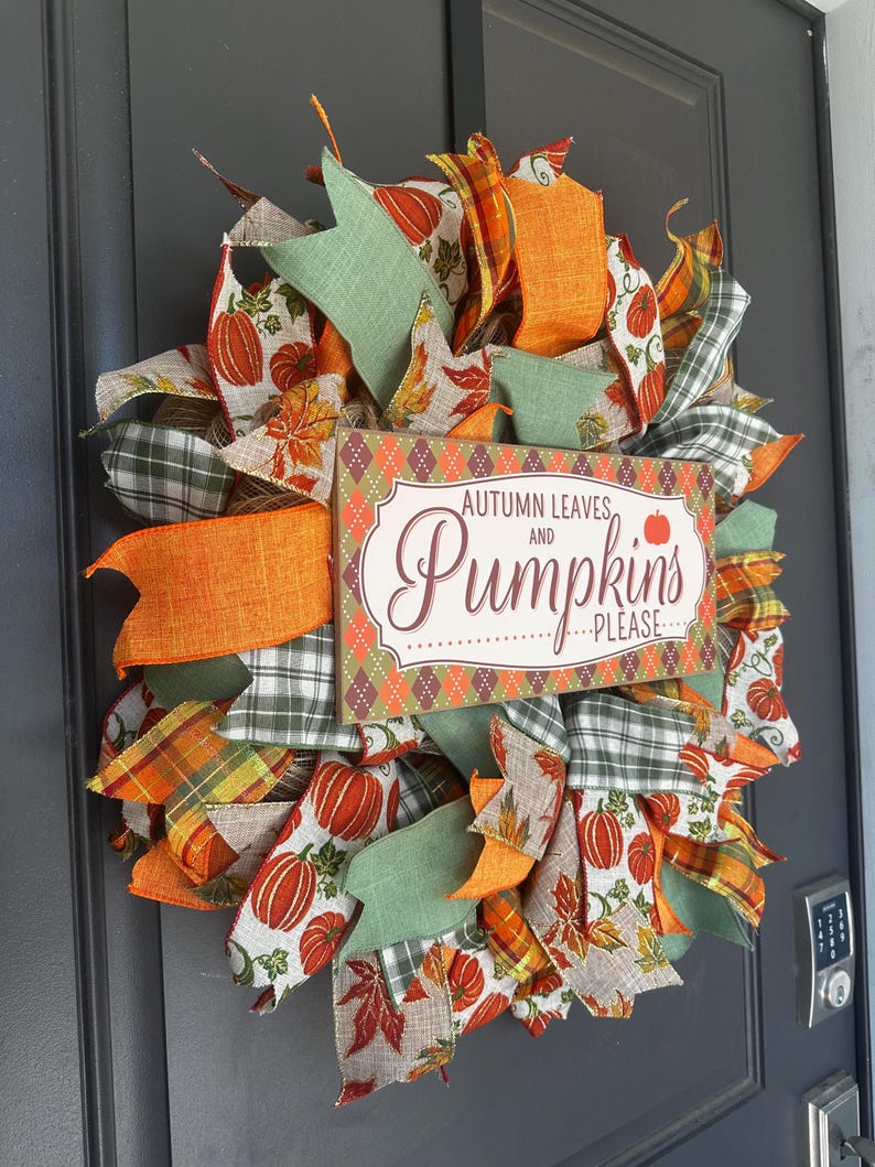 May include: A decorative wreath featuring orange, green, and white ribbons with pumpkin and leaf patterns. A rectangular wooden sign in the center displays the text Autumn Leaves and Pumpkins Please in brown and orange lettering on a cream background.