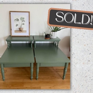 May include: Two green painted end tables with turned legs. The tables are stacked on top of each other. The top table has a framed print of a plant with a white background. The tables are sold.