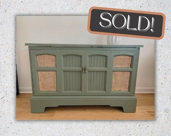 May include: A vintage green cabinet with a wood grain finish and two doors. The doors have a woven fabric panel on each side. The cabinet has a raised base and is sitting on a wood floor. The text "SOLD!" is written on a black chalkboard.