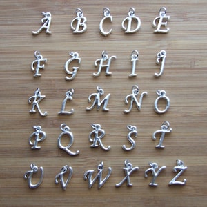 May include: Silver-colored alphabet charms, A through Z, displayed on a wooden surface. Each charm is a different letter in a cursive font, with a small loop at the top for attaching to jewelry or other crafts.