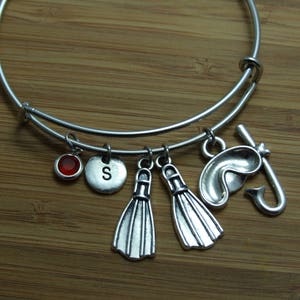 Op de afbeelding: Zilverkleurige, uitrekbare armband met bedels. Bedels zijn onder meer een rode edelsteen, een schijf met de letter "S", twee zwemvliezen en een snorkel met een masker. De armband ligt op een houten oppervlak.