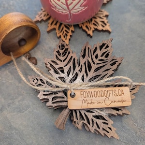 K&ouml;nnte beinhalten: Blattf&ouml;rmige Untersetzer aus Holz mit aufwendigen Ausschnitten. Ein Etikett lautet "FOXWOODGIFTS.CA Made in Canada". Eine rote Keramiktasse mit einem Blattmuster steht auf einem passenden Untersetzer. Eine Holzschaufel ist ebenfalls vorhanden.