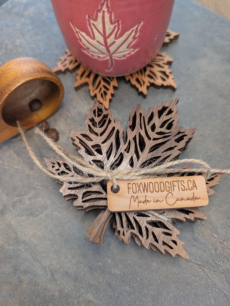 K&ouml;nnte beinhalten: Blattf&ouml;rmige Untersetzer aus Holz mit aufwendigen Ausschnitten. Ein Etikett lautet "FOXWOODGIFTS.CA Made in Canada". Eine rote Keramiktasse mit einem Blattmuster steht auf einem passenden Untersetzer. Eine Holzschaufel ist ebenfalls vorhanden.