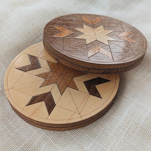 May include: Two circular wooden coasters featuring a geometric star pattern. The top coaster is made of dark brown wood with lighter wood accents, while the bottom coaster is made of light tan wood with dark brown wood geometric star inlays.