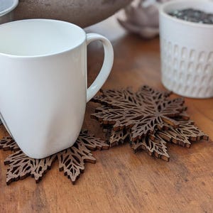 K&ouml;nnte beinhalten: Eine wei&szlig;e Keramiktasse steht auf einem braunen, lasergeschnittenen Holzuntersetzer in Ahornblattform. Zus&auml;tzliche Untersetzer sind daneben gestapelt. Die Tasse steht auf einer Holzoberfl&auml;che, im Hintergrund ein Metalleimer und ein wei&szlig;er Keramiktopf.