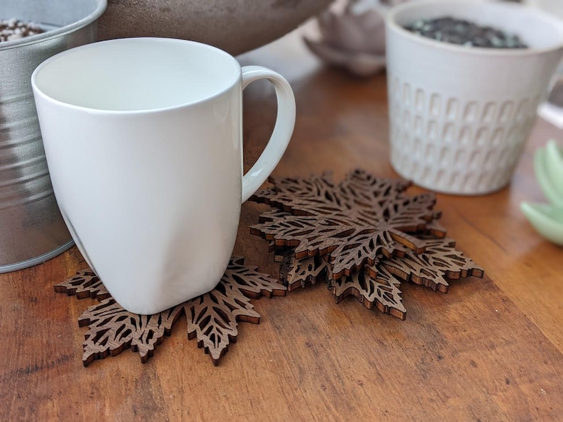 K&ouml;nnte beinhalten: Eine wei&szlig;e Keramiktasse steht auf einem braunen, lasergeschnittenen Holzuntersetzer in Ahornblattform. Zus&auml;tzliche Untersetzer sind daneben gestapelt. Die Tasse steht auf einer Holzoberfl&auml;che, im Hintergrund ein Metalleimer und ein wei&szlig;er Keramiktopf.