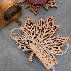 K&ouml;nnte beinhalten: Ein Stapel lasergeschnittener Ahornblatt-Untersetzer aus Holz, mit Schnur gebunden, mit einem Etikett mit der Aufschrift "FOXWOODGIFTS.CA Made in Canada". Eine rote Tasse mit Blattmuster steht auf einem passenden Untersetzer.