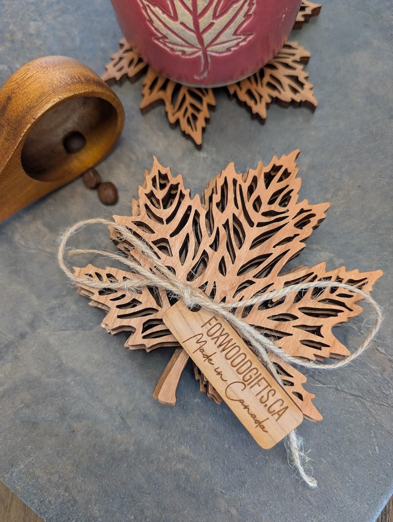 K&ouml;nnte beinhalten: Ein Stapel lasergeschnittener Ahornblatt-Untersetzer aus Holz, mit Schnur gebunden, mit einem Etikett mit der Aufschrift "FOXWOODGIFTS.CA Made in Canada". Eine rote Tasse mit Blattmuster steht auf einem passenden Untersetzer.