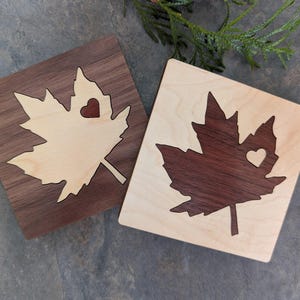 Peut inclure: Deux dessous de verre carrés en bois. L'un présente une feuille d'érable claire incrustée sur un fond en noyer foncé. L'autre a une feuille de noyer foncé incrustée sur un fond en érable clair. Chaque feuille a une découpe en forme de cœur.