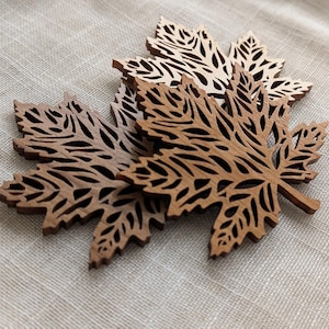Può includere: Diversi ritagli di foglie d'acero in legno in varie tonalità di marrone. Le foglie presentano intricati disegni traforati, che mostrano la venatura naturale del legno. Le foglie sono disposte in modo leggermente sovrapposto.