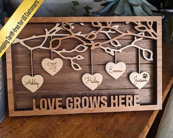 May include: A rectangular wooden wall sign featuring a layered tree branch design with five hanging heart-shaped ornaments. Each heart is engraved with a name: Ashley, Tj, Hailey, Emma, and Jaxson. The text LOVE GROWS HERE is at the bottom.