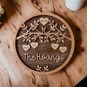 May include: A round wooden sign with a carved tree and hanging hearts. The hearts are inscribed with names, including "Adam", "Ruth", "Kai", and "Lyla". The sign also includes the text "The Hoangs" and "est. 2020".