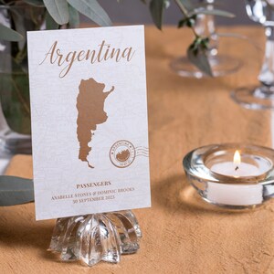 May include: A white card with a brown outline of Argentina and the text "Argentina" in brown cursive. The card also has a brown stamp with the text "Argentina" and a date. The card is on a silver stand and says "Passengers Anabelle Stones & Dominic Brooks 30 September 2023".