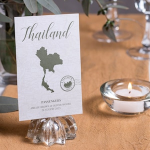 May include: A white card with a green outline of Thailand and the text "Thailand" in a cursive font. The card also includes a stamp with the text "Thailand" and a small image of a temple. The card is on a glass stand and says "Passengers Amelie Brown & Oliver Moore 26 August 2023".