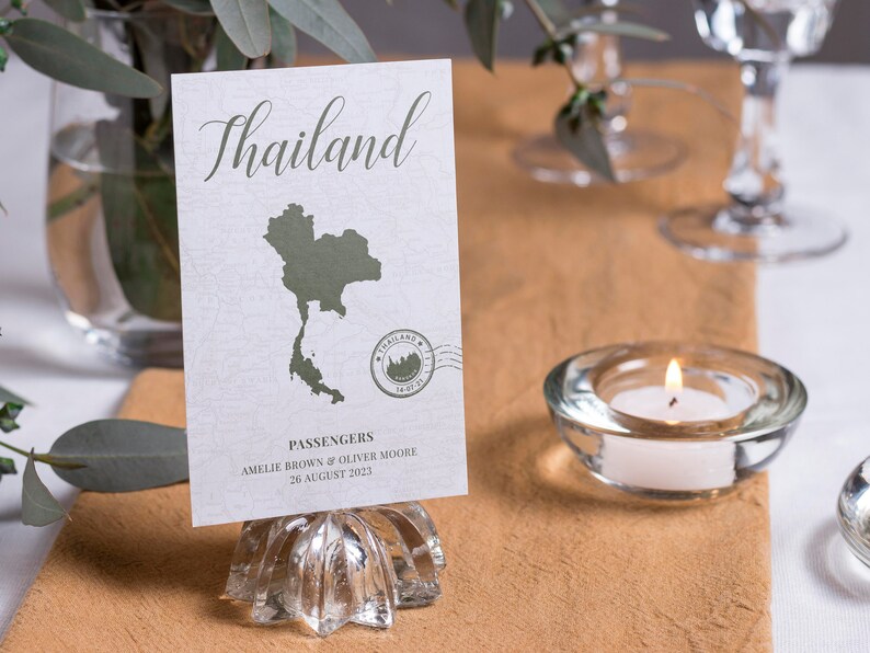 May include: A white card with a green outline of Thailand and the text "Thailand" in a cursive font. The card also includes a stamp with the text "Thailand" and a small image of a temple. The card is on a glass stand and says "Passengers Amelie Brown & Oliver Moore 26 August 2023".