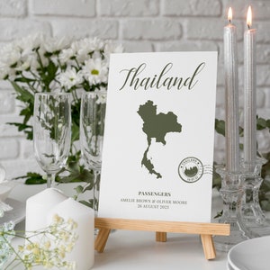 May include: A white card with the word "Thailand" written in cursive on it. The card also features a green outline of the country of Thailand and a stamp that says "Thailand 16-07-21". The card is on a wooden easel and there are two silver candlesticks with candles in them on the table.