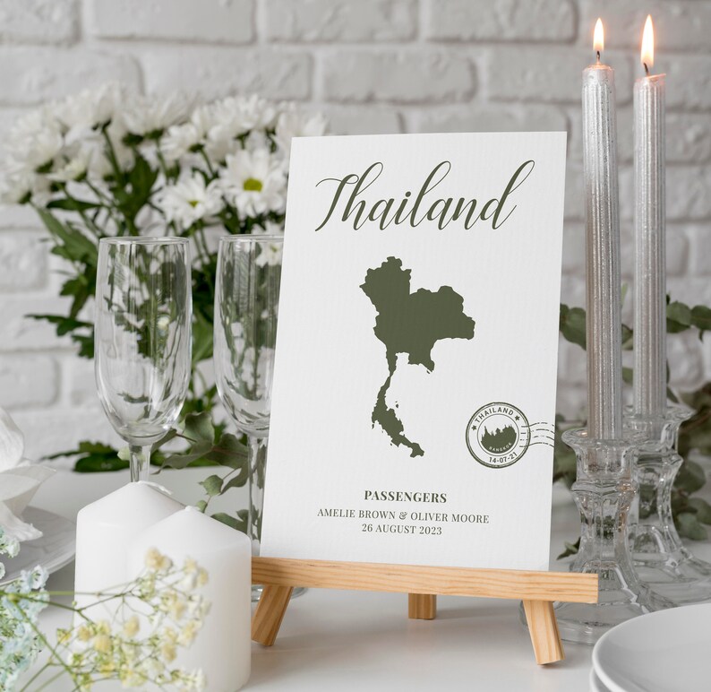 May include: A white card with the word "Thailand" written in cursive on it. The card also features a green outline of the country of Thailand and a stamp that says "Thailand 16-07-21". The card is on a wooden easel and there are two silver candlesticks with candles in them on the table.