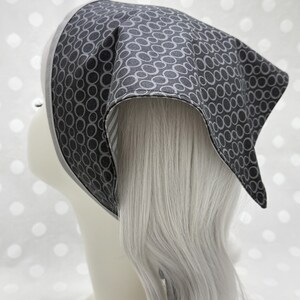 Puede incluir: Un pañuelo para la cabeza gris estilo bandana con un patrón ovalado repetido en un gris más oscuro. Los bordes están ribeteados con una tela gris más clara. El pañuelo se muestra en una cabeza de maniquí con pelo largo y plateado.