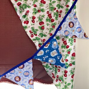 May include: A set of three blue and white triangle-shaped fabric banners with images of characters from the movie Frozen. The banners are strung together with blue ribbon and are hanging from a white fabric with a red strawberry pattern.