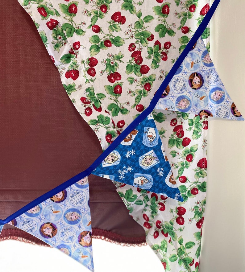 May include: A set of three blue and white triangle-shaped fabric banners with images of characters from the movie Frozen. The banners are strung together with blue ribbon and are hanging from a white fabric with a red strawberry pattern.