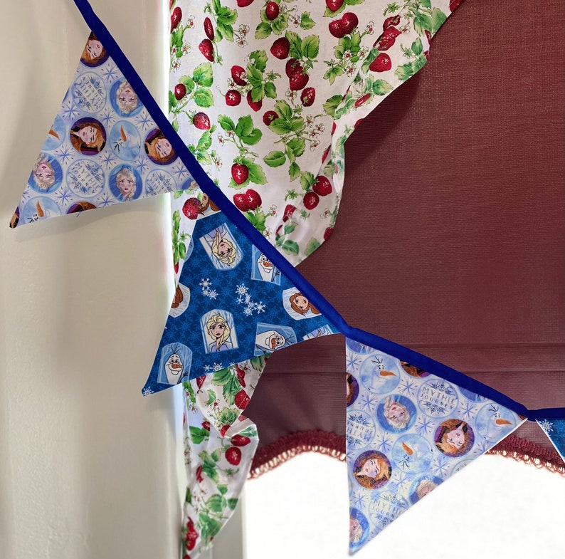 May include: A blue and white fabric banner with images of characters from the movie Frozen. The banner is decorated with blue and white ribbon and is hanging from a white wall.