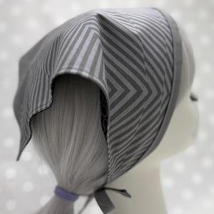 Puede incluir: Un pañuelo gris con un diseño de chevron y rayas. El cubrecabezas está atado en la nuca con un pequeño lazo. La tela es gris claro con rayas grises más oscuras.