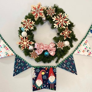 Op de afbeelding: Een kerstkrans met witte en rode sneeuwvlokken en een rood-witte strik. Een banner met de woorden "Merry Merry" en "La La La" hangt onder de krans.