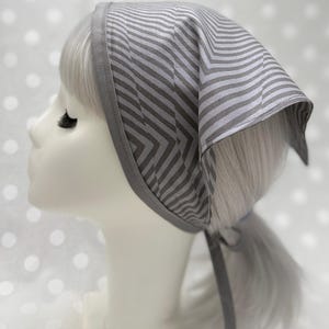 Puede incluir: Un pañuelo para la cabeza a rayas grises y blancas con un diseño de chevrón. El cubrecabezas se ata en la nuca con lazos grises a juego. El pañuelo tiene un borde gris liso.
