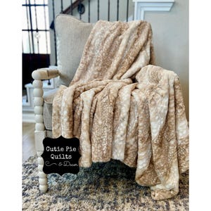May include: A soft, beige faux fur throw blanket with a subtle pattern draped over a white chair. The chair has a wooden base with decorative round knobs. A black sign with white text reads "Cutie Pie Quilts & Decor".