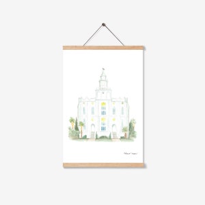 May include: Watercolor print of a white building with a steeple and cross, likely a temple. The artwork is framed by a wooden magnetic poster hanger. Palm trees and greenery surround the building. Signed by the artist, Rebecca Ann.
