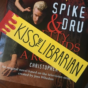 May include: A yellow bookmark with the text "Kiss the Librarian" in black lettering. The bookmark is partially covering a book titled "Spike & Dru" with a red and black background.