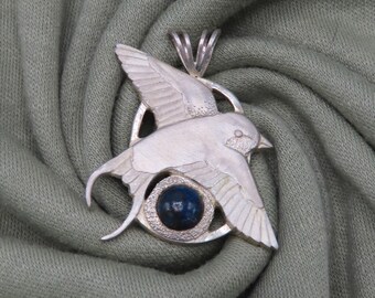 May include: A silver pendant featuring a stylized bird with a blue gemstone eye. The pendant is on a green fabric background.