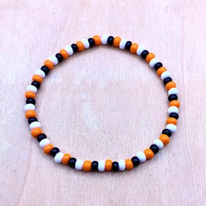 May include: A black, white, and orange beaded bracelet. The bracelet is made of small, round beads that are strung together in a repeating pattern.