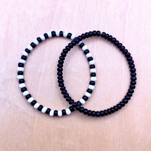 May include: Two beaded bracelets, one black and one black and white, are intertwined. The bracelets are made of small round beads.