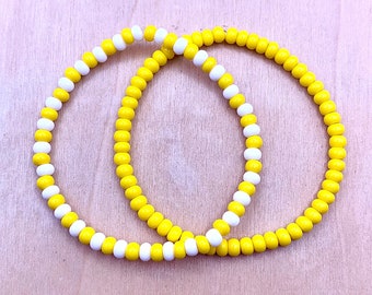 May include: Two yellow and white beaded bracelets. The bracelets are made of small, round beads and are stacked on top of each other.