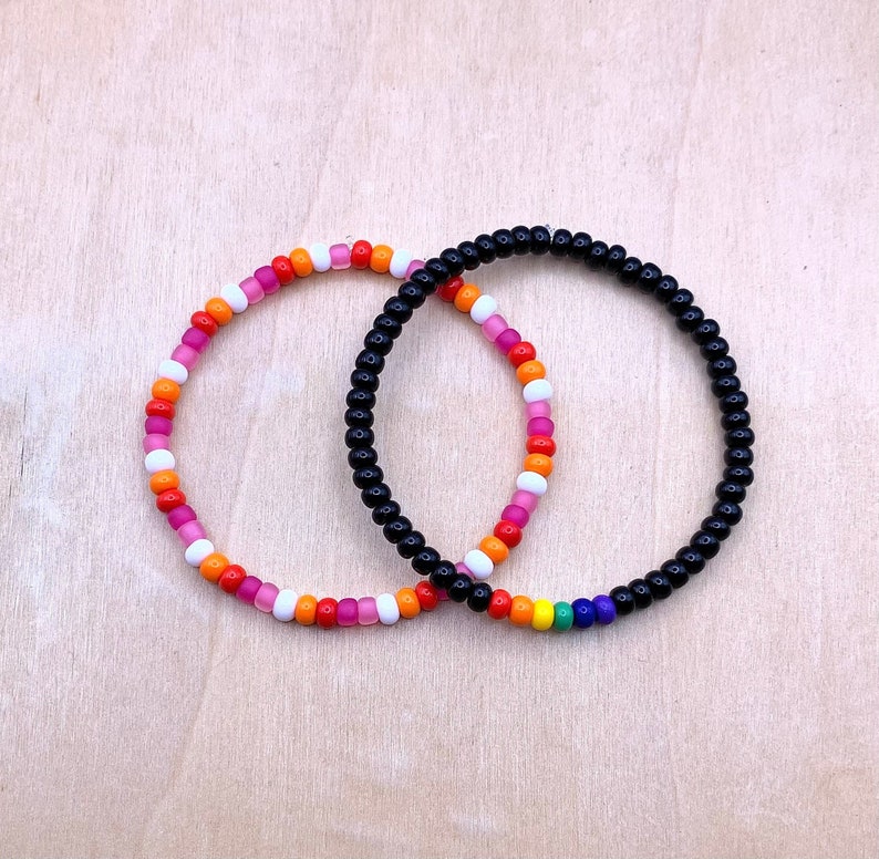 Puede incluir: Dos pulseras de cuentas, una con cuentas rosas, blancas y naranjas, y la otra con cuentas negras y una franja de arco&iacute;ris.