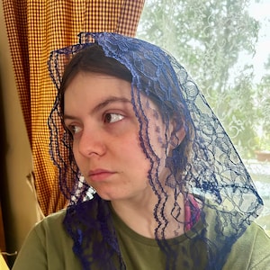 May include: A woman wearing a blue lace head covering. The lace is delicate and has a scalloped edge. The woman is looking to the side and has a serious expression on her face.